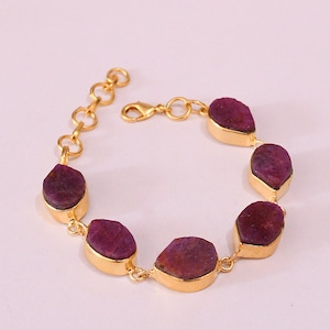 Peut inclure: Un bracelet doré orné de sept pierres précieuses rouge foncé de forme irrégulière. Chaque pierre est sertie dans une lunette dorée et reliée par des maillons dorés, avec un fermoir mousqueton. Le bracelet est présenté sur un fond clair.