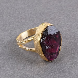 Op de afbeelding: Een goudkleurige ring met een grote, ruw gesneden, dieprode edelsteen. De edelsteen is gezet in een decoratieve bezel met geëtste bloempatronen. De ringband heeft een textuurontwerp.