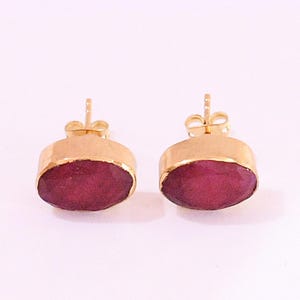 May include: A pair of stud earrings featuring oval-shaped, deep red faceted stones set in gold-toned metal bezels. Each earring includes a standard post back mechanism for secure attachment, showcasing a polished finish on the metal edges.