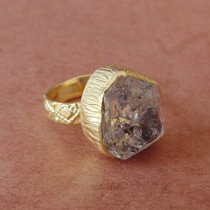 Puede incluir: Un anillo dorado con un cristal grande, claro y de forma irregular. El anillo tiene una banda texturizada y un engaste decorativo alrededor del cristal. El fondo es de un marrón rojizo apagado.