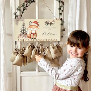 Puede incluir: Calendario de Adviento de madera con una ilustración de un zorro con gorro de Papá Noel, sosteniendo un regalo. El calendario tiene pequeñas bolsas de arpillera colgando debajo, cada una con un número. El texto "Adventskalender von Petra" está escrito en la tabla de madera. Una niña sonríe.