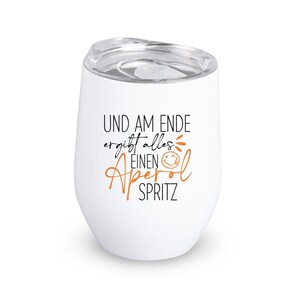 May include: White stemless wine tumbler with a clear lid. The tumbler has the text "UND AM ENDE ergibt alles EINEN Aperol SPRITZ" in black and orange script. The design includes a smiling face.