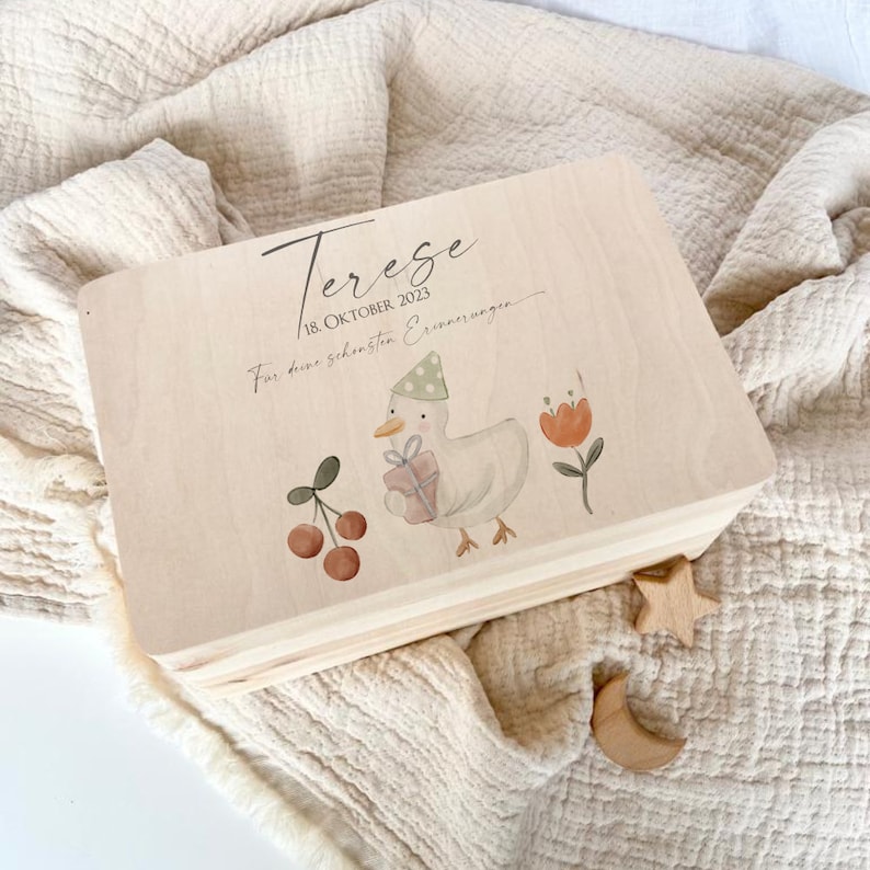May include: A wooden keepsake box with a light wood finish. The box has a painted design of a duck holding a gift, cherries, and flowers. The text on the box reads "Terezse 18. Oktober 2023 F&uuml;r deine sch&ouml;nsten Erinnerungen."