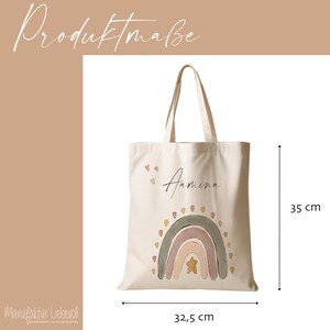 Peut inclure: Un sac fourre-tout en toile blanche avec un motif arc-en-ciel dans des tons de rose, vert et beige. Le sac porte le nom "Amina" &eacute;crit en cursive sur le devant. Le sac mesure 32,5 cm de large et 35 cm de haut.