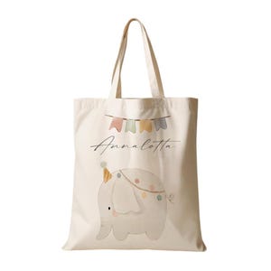 Peut inclure: Un sac fourre-tout en toile beige avec un &eacute;l&eacute;phant gris portant un chapeau de f&ecirc;te et une guirlande de fanions color&eacute;s. Le sac porte le nom "Annalotta" imprim&eacute; dessus.