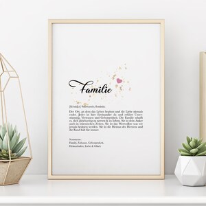 Puede incluir: Un diseño de arte de pared imprimible con un fondo blanco y dorado y un corazón rosa. La palabra "Familie" está escrita en cursiva negra. El texto de abajo define la palabra "Familie" en alemán, explicando su significado como un lugar de amor, apoyo y pertenencia.