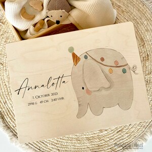 May include: A wooden keepsake box with a light wood finish. The box features a cute illustration of a grey elephant wearing a party hat and a colourful garland. The text "Annalotta" is written in cursive on the box, along with the date "7. Oktober 2023", weight "2998 G", height "49 CM", and time "2:40 UHR".