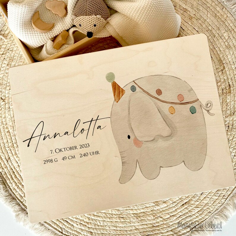 May include: A wooden keepsake box with a light wood finish. The box features a cute illustration of a grey elephant wearing a party hat and a colourful garland. The text "Annalotta" is written in cursive on the box, along with the date "7. Oktober 2023", weight "2998 G", height "49 CM", and time "2:40 UHR".