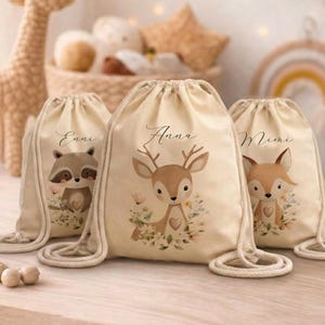 Puede incluir: Tres mochilas de cordón en color blanco roto hechas de tela de lona. Cada bolsa presenta una ilustración animal única: un mapache etiquetado como Enni, un ciervo como Anna y un zorro como Mimi. Cada animal está rodeado de detalles florales.