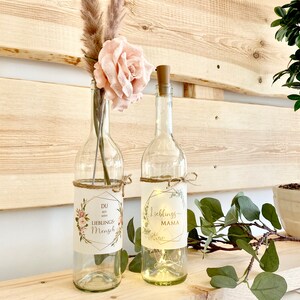 May include: Two glass bottles with cork tops decorated with floral and greenery designs. The bottle on the left has a label that reads "Du bist mein Lieblings Mensch" and the bottle on the right has a label that reads "Lieblings Mama".