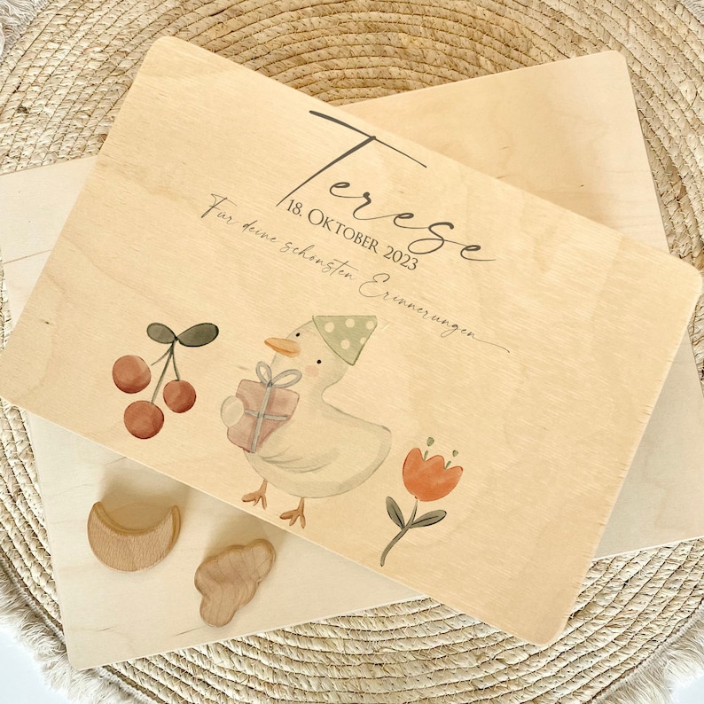 May include: A wooden box with a light wood finish. The box has a lid with a hand-painted design of a chick holding a gift, a cherry branch, and a red flower. The lid also has the text "Terezse 18 OKTOBER 2023 F&uuml;r deine sch&ouml;nsten Erinnerungen".