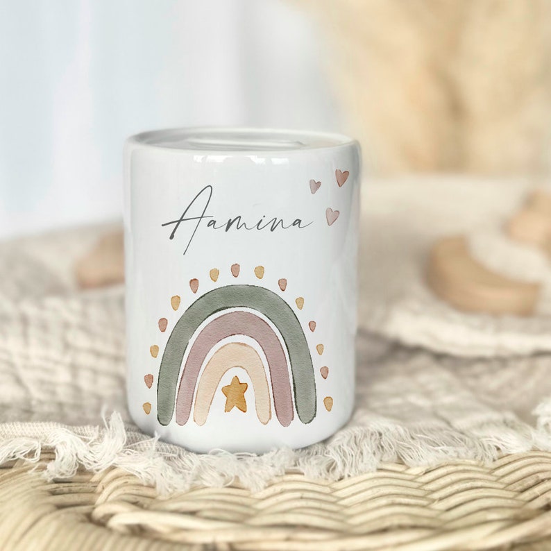 May include: White ceramic piggy bank with a rainbow design and the name "Amina" written on it. The rainbow is in shades of pink, peach, and green with a yellow star in the center.