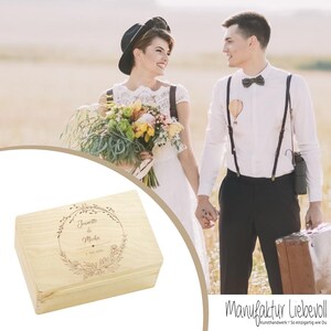 May include: A wooden box with an engraved floral design and the text "Janette & Micha 1. Mai 2023". The box is light brown and has a lid.