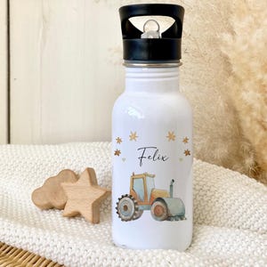 May include: White stainless steel water bottle with a black lid and a yellow tractor design. The bottle has a name, "Felix", written on it with gold stars.