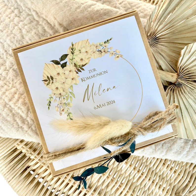 May include: A brown box with a white card inside. The card has a floral design with white flowers and a gold circle. The text on the card reads "Zur Kommunion Milena 6. Mai 2024".