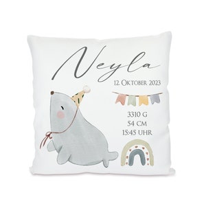 May include: A white pillow with a watercolor illustration of a seal wearing a party hat. The pillow also features a rainbow, bunting, and the text "Neyla 12. OKTOBER 2023 3310 G 54 CM 15:45 UHR".