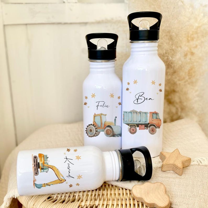 May include: Three white stainless steel water bottles with black lids and handles. Each bottle has a different design featuring a construction vehicle and a name: Felix, Ben, and Armand. The bottles are on a woven wicker surface with wooden stars.