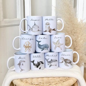 Puede incluir: Conjunto de 9 tazas de cerámica blanca con ilustraciones de animales marinos y nombres personalizados. Las tazas presentan un tiburón, una foca, un pulpo, una ballena, una estrella de mar, un delfín, una orca, un tiburón ballena y una tortuga marina.