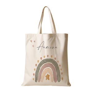 Peut inclure: Un sac fourre-tout en toile beige avec un motif arc-en-ciel dans des tons de vert, rose et beige. L'arc-en-ciel a une &eacute;toile jaune au centre. Le sac porte le nom "Amina" &eacute;crit en cursive.