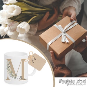 Puede incluir: Taza de cer&aacute;mica blanca con una letra dorada "M" y el nombre "Marie" impreso en ella. La taza est&aacute; decorada con un dise&ntilde;o floral verde y blanco. Una etiqueta de regalo marr&oacute;n con el texto "F&uuml;r  Von" est&aacute; unida a la taza.