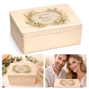 Op de afbeelding: Een lichtgekleurde houten kist met een scharnierend deksel. Het deksel heeft een ovaal bloemenontwerp met het woord "Name" in script. De kist is versierd met een botanische krans en de tekst "DEIN WUNSCHTEXT".