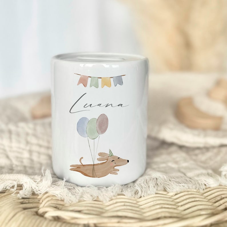 May include: White ceramic piggy bank with a cartoon dachshund wearing a party hat and holding balloons. The piggy bank has the name "Luana" written on it and colorful bunting above the name.