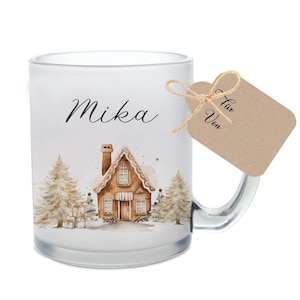 Könnte beinhalten: Eine weiße, gefrostete Glas-Tasse mit einem Design aus Lebkuchenhaus und schneebedeckten Bäumen. Die Tasse trägt den Namen "Mika" und ein braunes Geschenkanhänger mit den Worten "Für" und "Von" darauf.
