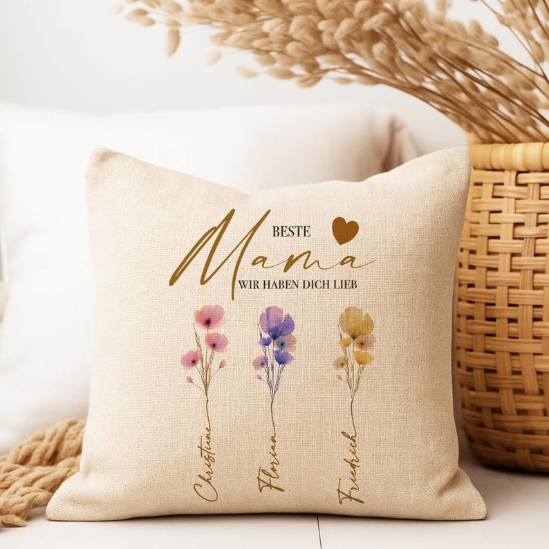 May include: Beige square throw pillow with the words "Beste Mama" and "Wir haben dich lieb" in gold script. The pillow features watercolor floral designs in pink, purple, and yellow, with names below each flower.