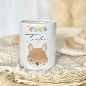 May include: White ceramic piggy bank with a fox illustration and the name "Triton" written in cursive. The bank has a colorful string of flags on top.