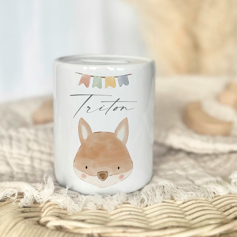 May include: White ceramic piggy bank with a fox illustration and the name "Triton" written in cursive. The bank has a colorful string of flags on top.