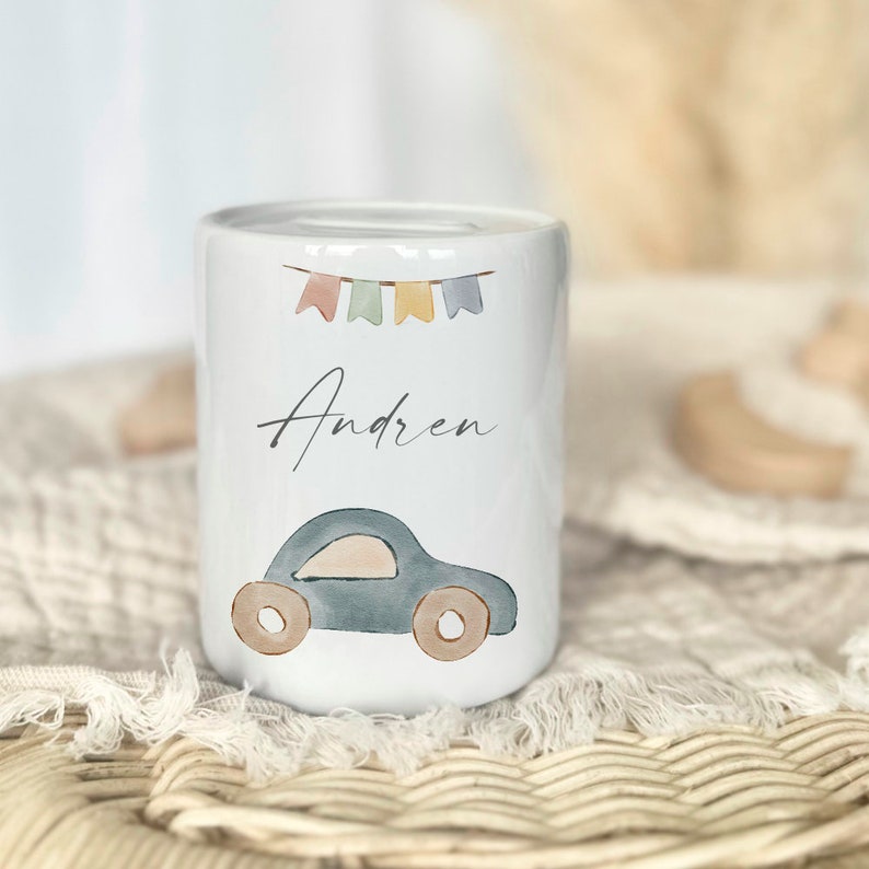 May include: White ceramic piggy bank with a blue car, colorful flags, and the name "Andren" written on it.