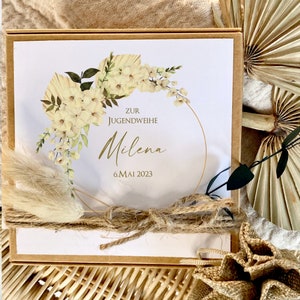 May include: A white card with a floral wreath design and gold lettering. The card says "Zur Jugendweihe Milena 6. Mai 2023". The card is decorated with a brown rope and a dried flower.