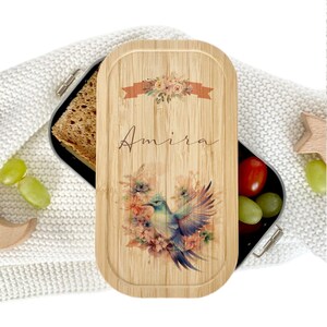 Könnte beinhalten: Eine personalisierte Bambus-Lunchbox mit einem Blumendesign und einem blauen Vogel. Der Name "Amira" ist auf dem Deckel geschrieben.
