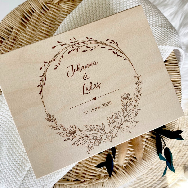 May include: A wooden box with a floral wreath design engraved on the lid. The wreath includes the names "Johanna & Lukas" and the date "10. JUNI 2023".