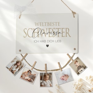 Può includere: Un cartello di legno bianco con il testo "Weltbeste Schwester Clarissa Ich hab dich lieb" in lettere nere e dorate. Un simbolo di cuore si trova sotto il testo. Uno spago di spago con mollette per bucato tiene cinque foto di bambini.