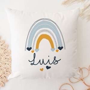 May include: A white pillow with a blue, yellow, and orange rainbow design. The rainbow has small hearts hanging from it. The name "Luis" is written in black below the rainbow.