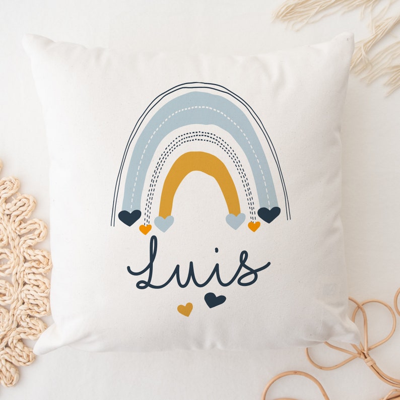 May include: A white pillow with a blue, yellow, and orange rainbow design. The rainbow has small hearts hanging from it. The name "Luis" is written in black below the rainbow.