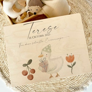 May include: A wooden keepsake box with a light wood finish. The box has a duck with a party hat holding a gift, cherries, and a flower. The text on the box reads "Tereze 18. OKTOBER 2023 F&uuml;r deine sch&ouml;nsten Erinnerungen".