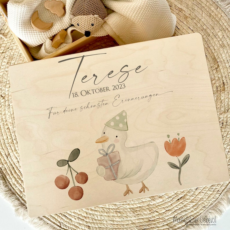 May include: A wooden keepsake box with a light wood finish. The box has a duck with a party hat holding a gift, cherries, and a flower. The text on the box reads "Tereze 18. OKTOBER 2023 F&uuml;r deine sch&ouml;nsten Erinnerungen".
