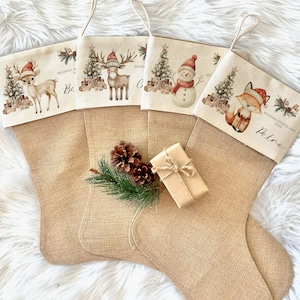 Può includere: Quattro calze natalizie in juta con disegni festivi. Ogni calza presenta un personaggio festivo diverso: un cervo, una renna, un pupazzo di neve e una volpe, tutti con cappelli di Babbo Natale. Un piccolo pacco regalo e pigne sono di fronte.