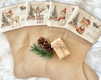 May include: Four burlap Christmas stockings with festive designs. Each stocking features a different holiday character: a deer, a reindeer, a snowman, and a fox, all wearing Santa hats. A small gift box and pinecones are in front of the stockings.