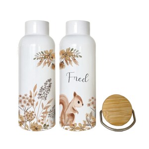 Könnte beinhalten: Zwei weiße Keramik-Wasserflaschen mit einem Aquarell-Design aus Blättern, Blumen und einem Eichhörnchen. Die Flasche rechts trägt den Namen "Fred". Die Flaschen haben Bambusdeckel mit einem Metallring.