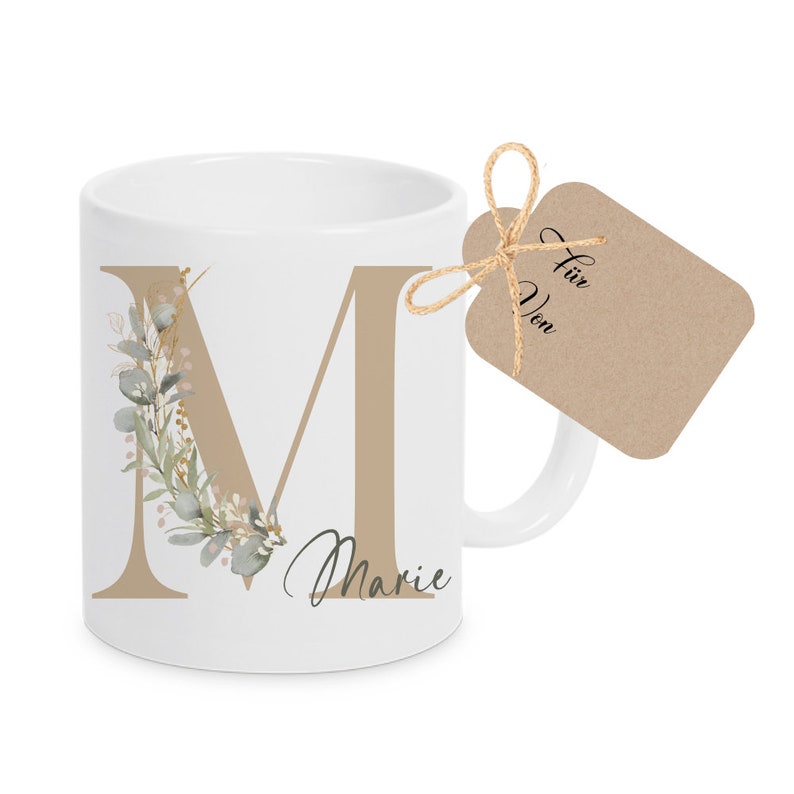 Puede incluir: Taza de cer&aacute;mica blanca con una gran letra "M" marr&oacute;n decorada con un dise&ntilde;o floral verde y marr&oacute;n. La taza tiene el nombre "Marie" escrito debajo de la letra. Una etiqueta de regalo marr&oacute;n con el texto "F&uuml;r Von" est&aacute; atada a la taza con un cordel.
