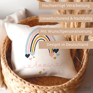 May include: A white pillow with a rainbow design and the name "emmare" in pink script. The pillow is in a wicker basket. The text "Hochwertige Verarbeitung", "Umweltschonend & Nachhaltig", "Mit Wunschpersonalisierung", "Designt in Deutschland", and "Manufaktur Liebevoll" is visible in the image.