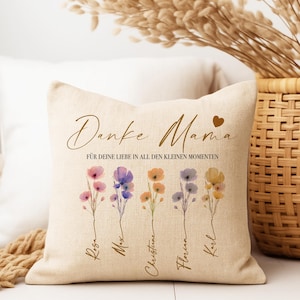 May include: Beige square throw pillow with floral design and text. The pillow features watercolor flowers in pink, purple, orange, and yellow. The text reads "Danke Mama" and "F&Uuml;R DEINE LIEBE IN ALL DEN KLEINEN MOMENTEN" with names below the flowers.