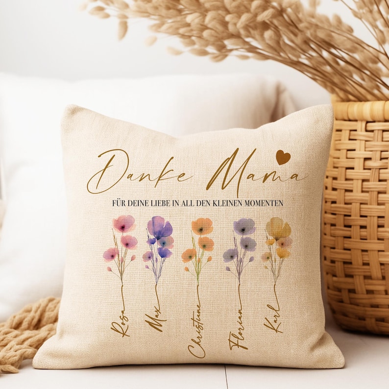 May include: Beige square throw pillow with floral design and text. The pillow features watercolor flowers in pink, purple, orange, and yellow. The text reads "Danke Mama" and "F&Uuml;R DEINE LIEBE IN ALL DEN KLEINEN MOMENTEN" with names below the flowers.