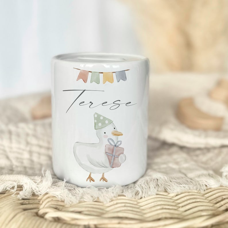 May include: White ceramic piggy bank with a cartoon duck wearing a party hat and holding a gift. The bank has the name "Terese" written on it and colorful flags above the name.