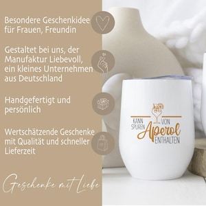 May include: A white stemless wine tumbler with a silver lid. The tumbler has the words "Kann Spuren von Aperol Enthalten" in gold script and a cocktail illustration. The background includes a beige vase and text in German.
