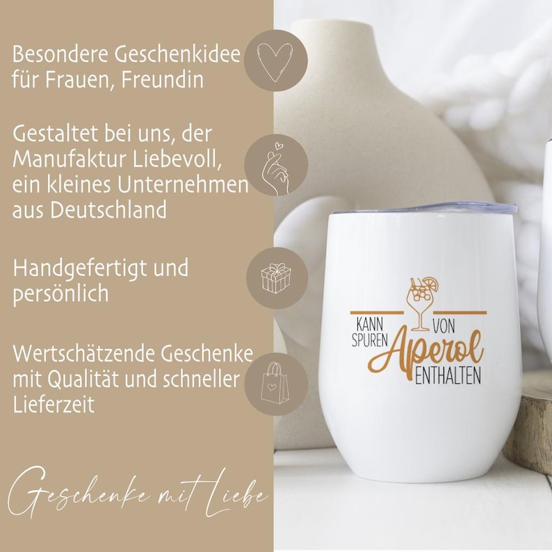 May include: A white stemless wine tumbler with a silver lid. The tumbler has the words "Kann Spuren von Aperol Enthalten" in gold script and a cocktail illustration. The background includes a beige vase and text in German.