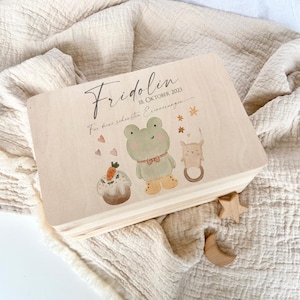 Peut inclure: Une boîte à souvenirs en bois avec une finition en bois clair. La boîte est décorée d'un motif peint d'une grenouille, d'un lapin et d'un gâteau avec une carotte sur le dessus. Le texte sur la boîte est "Fridolin 18. Oktober 2023 Für deine schönsten Erinnerungen".